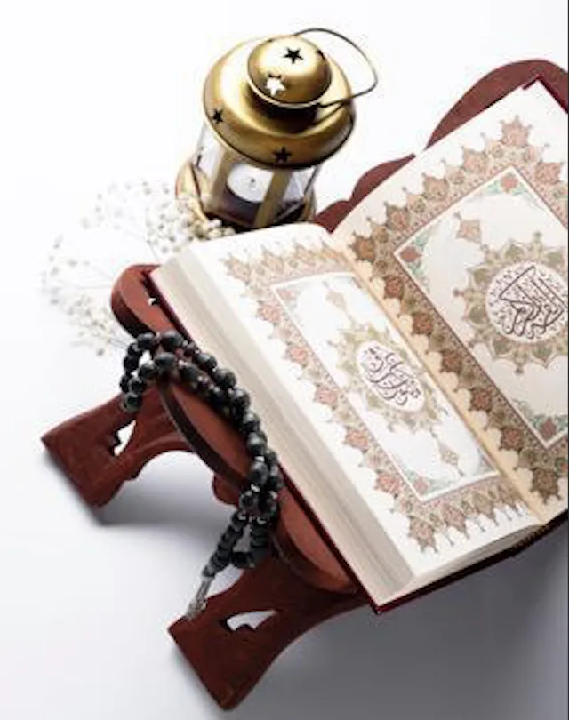 An open Quran on a wooden rehl beside a lantern and prayer beads